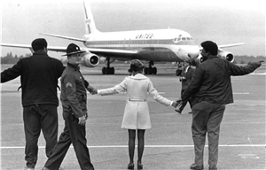 A small woman in a white coat stands between two large men in black coats. They are holding hands and facing an airplane on the runway. A police officer walks by and looks at the camera. 